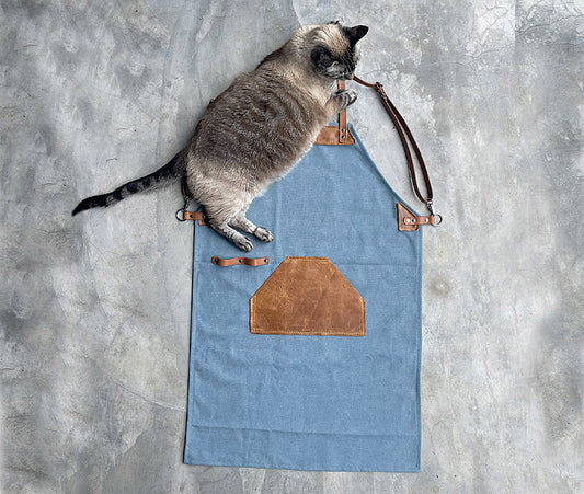 Handmade blue canvas apron with brown leather straps and pocket, designed for durability and style, perfect for chefs, artists, and craftsmen. Adjustable leather straps for a comfortable fit and a large leather pocket for tools and essentials.