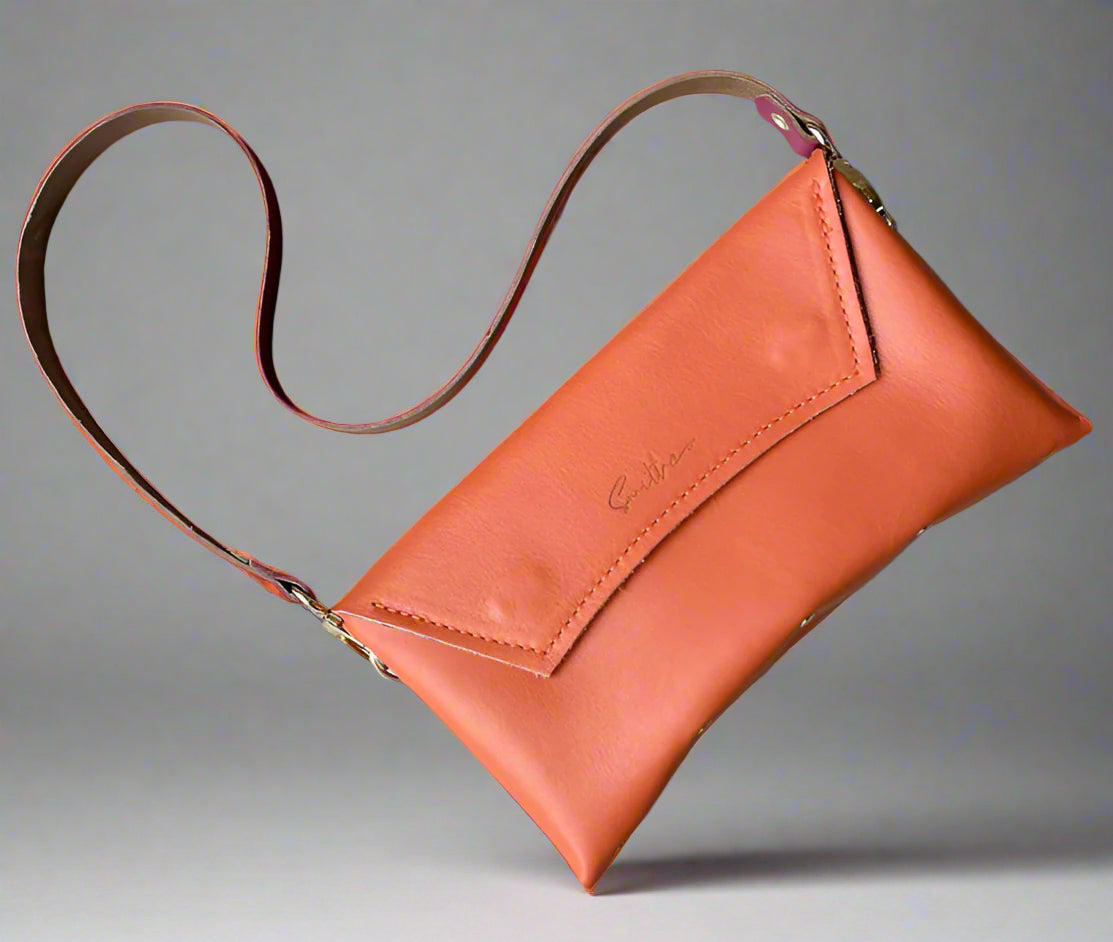 An orange full grain leather clutch sized bag with rose pink coloured detail, stunning curved lines, and studs along the base.
