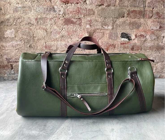 Side view of a full grain leather duffel bag in a dark forest green with brown details pictured against a concrete screed floor and rough red brick wall.