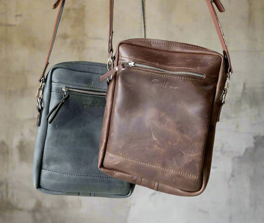 Full-grain leather crossbody sling bag in distressed brown and denim blue, featuring an two exterior zipped pockets and adjustable strap against a rustic wall backdrop.